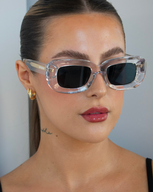 Close-up image of a female with an oval face shape wearing our limited edition Unfazed sunglasses, handcrafted in Italy for a universal fit. The sunglasses feature a transparent thick frame and grey see-through lens.