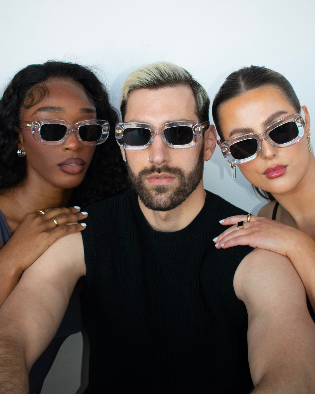 Image featuring three individuals wearing Rewrites Unfazed Sunglasses with thick transparent frames and grey see-through lenses. The model on the left, Nyallay, has an oval face shape. In the middle, male model Max also has an oval face shape. On the right, another model with a round face shape is depicted.
