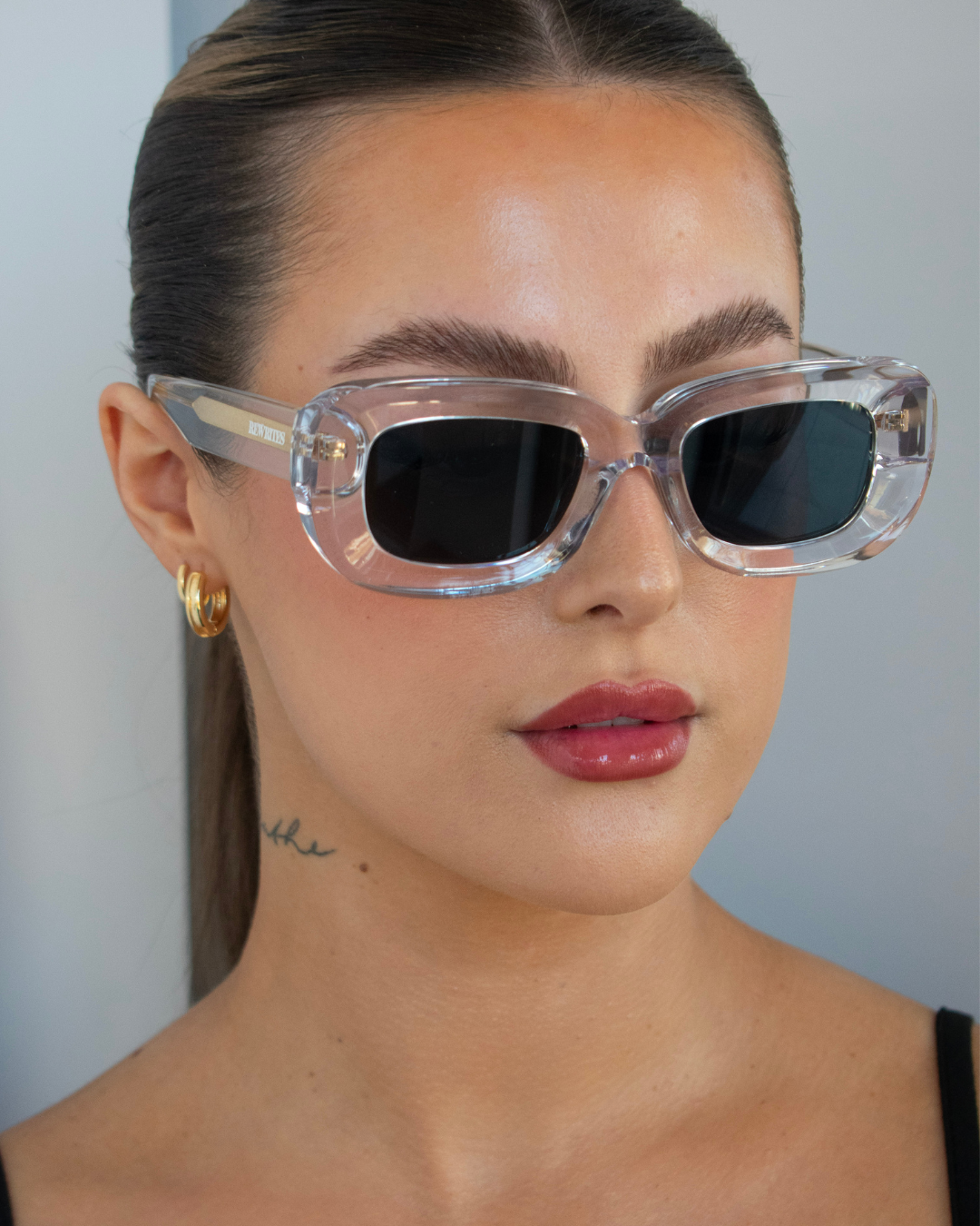 Close-up image of a female with an oval face shape wearing our limited edition Unfazed sunglasses, handcrafted in Italy for a universal fit. The sunglasses feature a transparent thick frame and grey see-through lens.